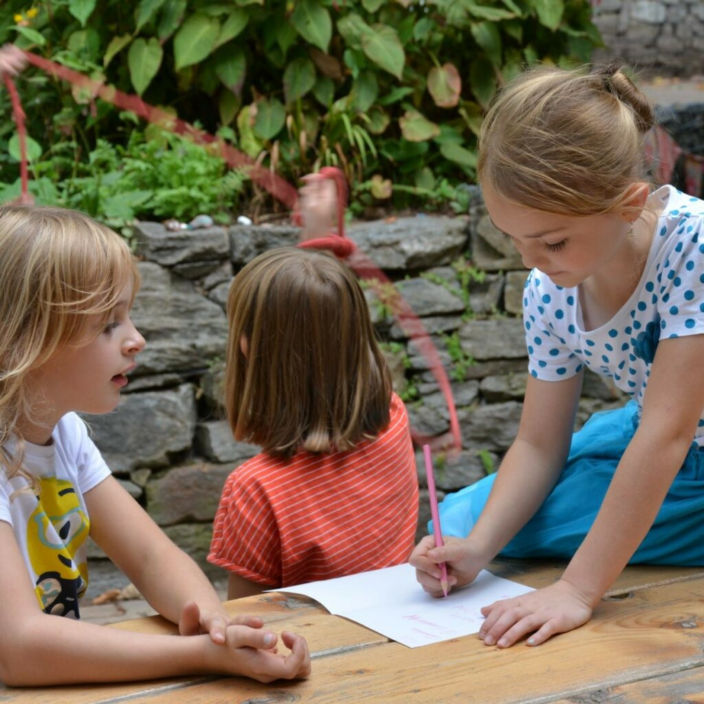 Drei Kinder sitzen draußen an einem Holztisch, ein Mädchen malt auf einem Blatt Papier.